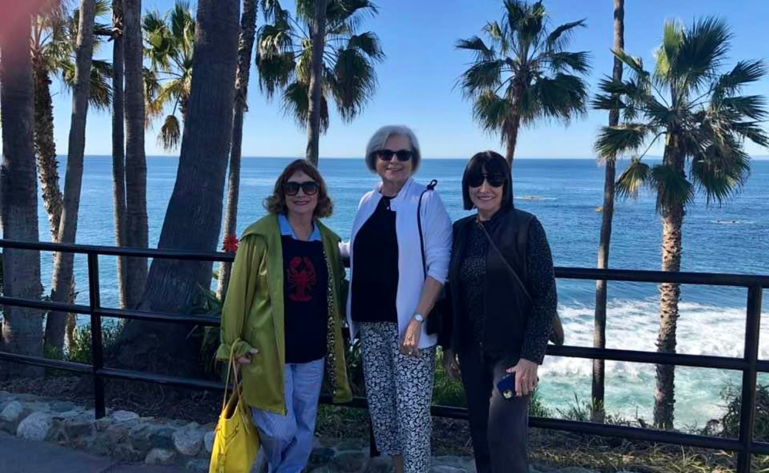 Three women posing together near palm trees and the ocean during a mother-daughter getaway on the California coast.