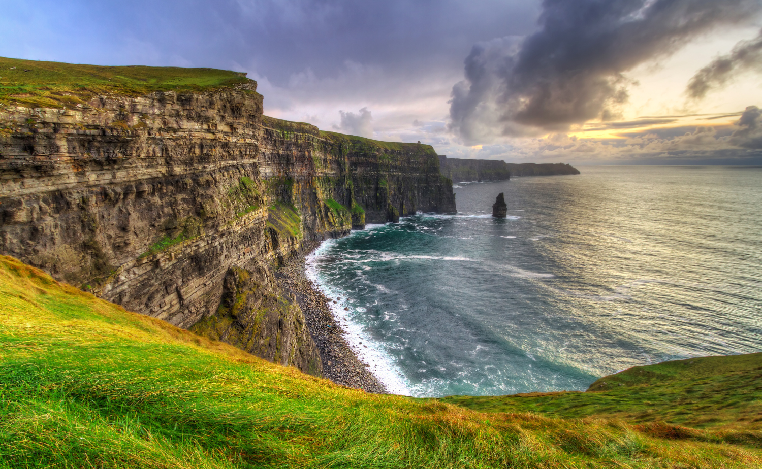 A dramatic coastal view of the Cliffs of Moher with rugged cliffs, green grass, and waves crashing along the Atlantic Ocean.