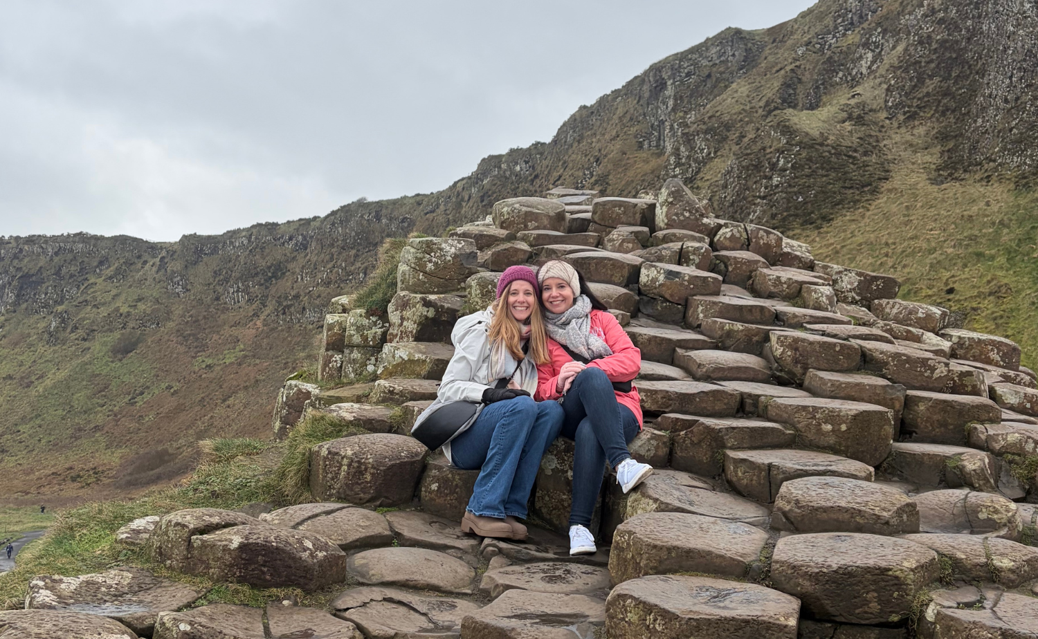 Two travelers sitting on the hexagonal basalt columns at Giant’s Causeway along Northern Ireland’s dramatic Antrim Coast under a cloudy sky.