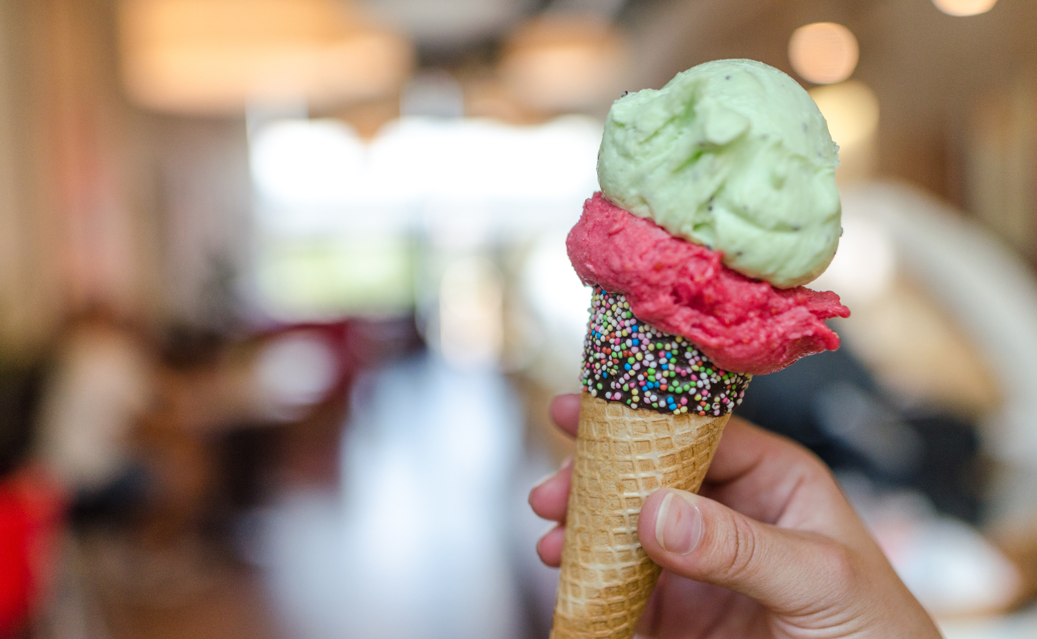 A hand holding a cone of pistachio and strawberry gelato inside a gelato shop in Rome.