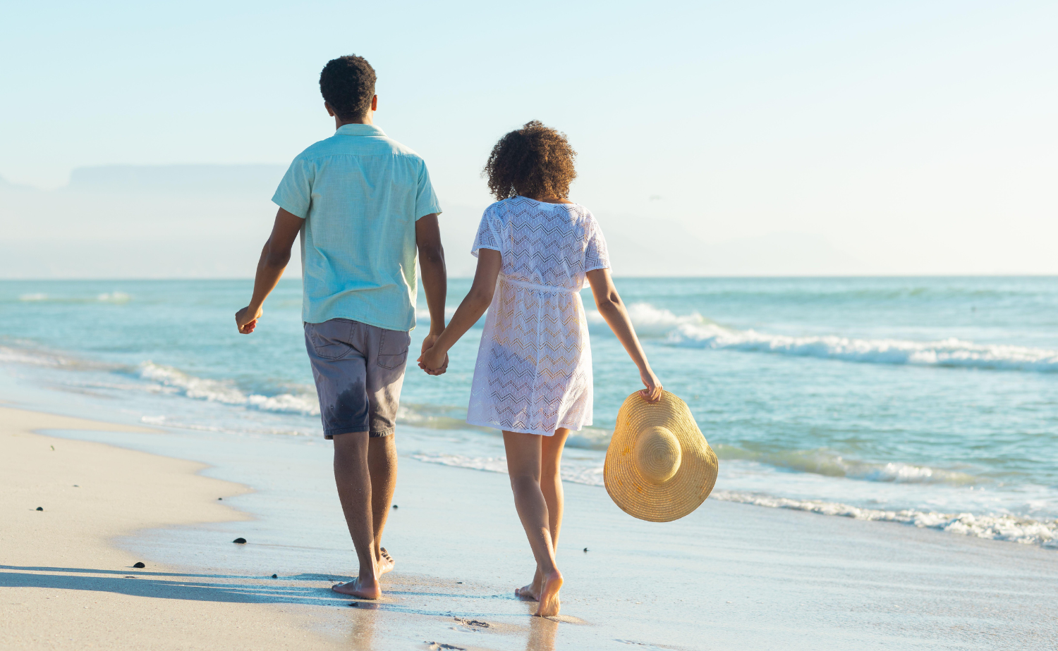 A couple walks hand in hand along a peaceful shoreline with waves rolling in.