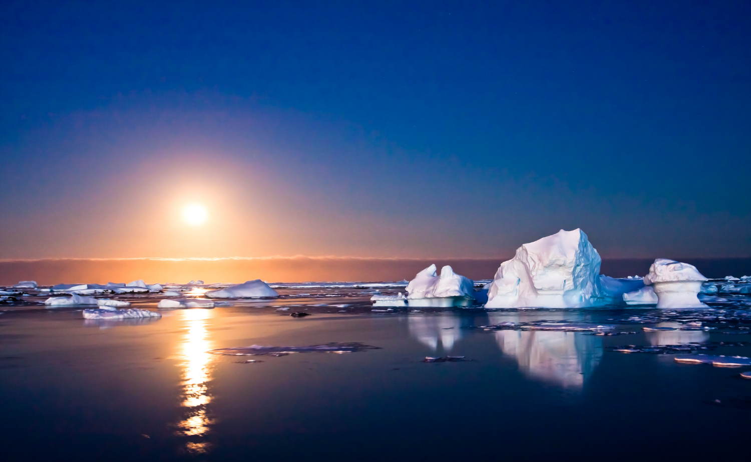 Icebergs floating in calm Antarctic water as the sun sets over the horizon