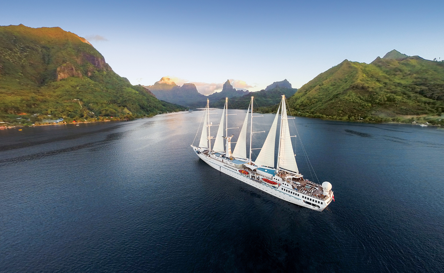 A Windstar sailing yacht cruises through calm waters surrounded by lush, green peaks in Moorea at sunset.