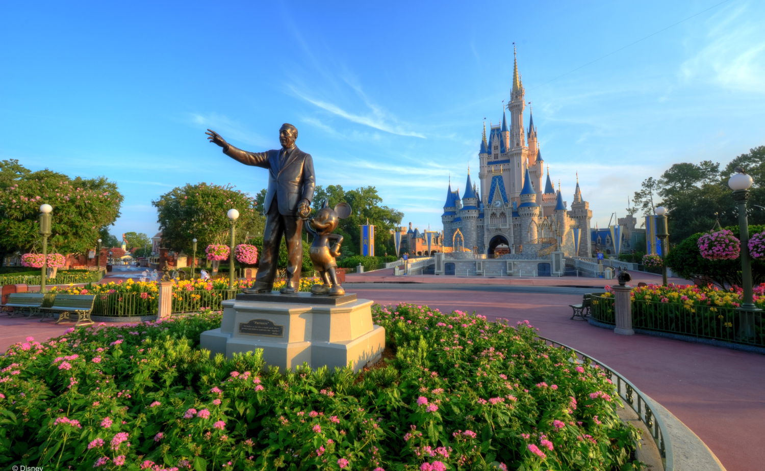 Statue of Walt Disney and Mickey Mouse with Cinderella Castle in the background at Magic Kingdom, surrounded by colorful flowers.