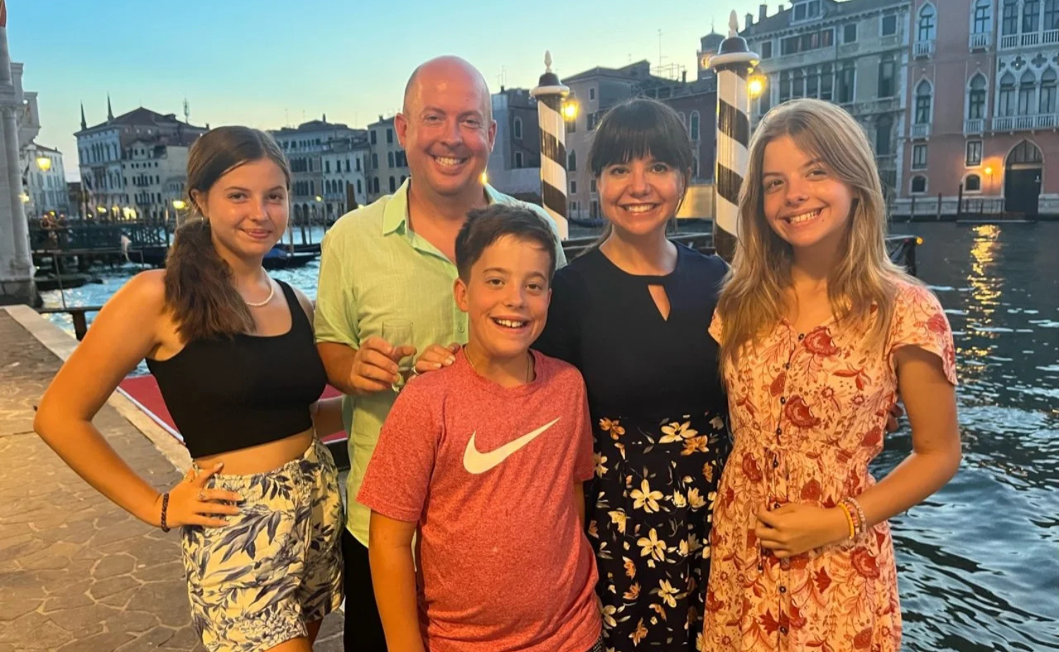 A family of five smiles together along a canal in Venice at sunset, with historic buildings and water behind them.