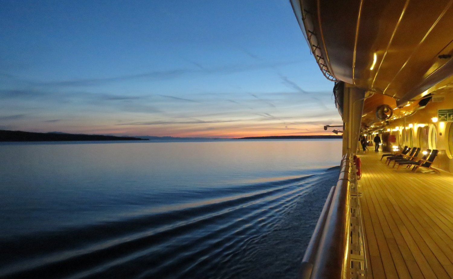 A cruise ship deck glows under warm lights as it sails into an Alaskan sunset over calm waters.