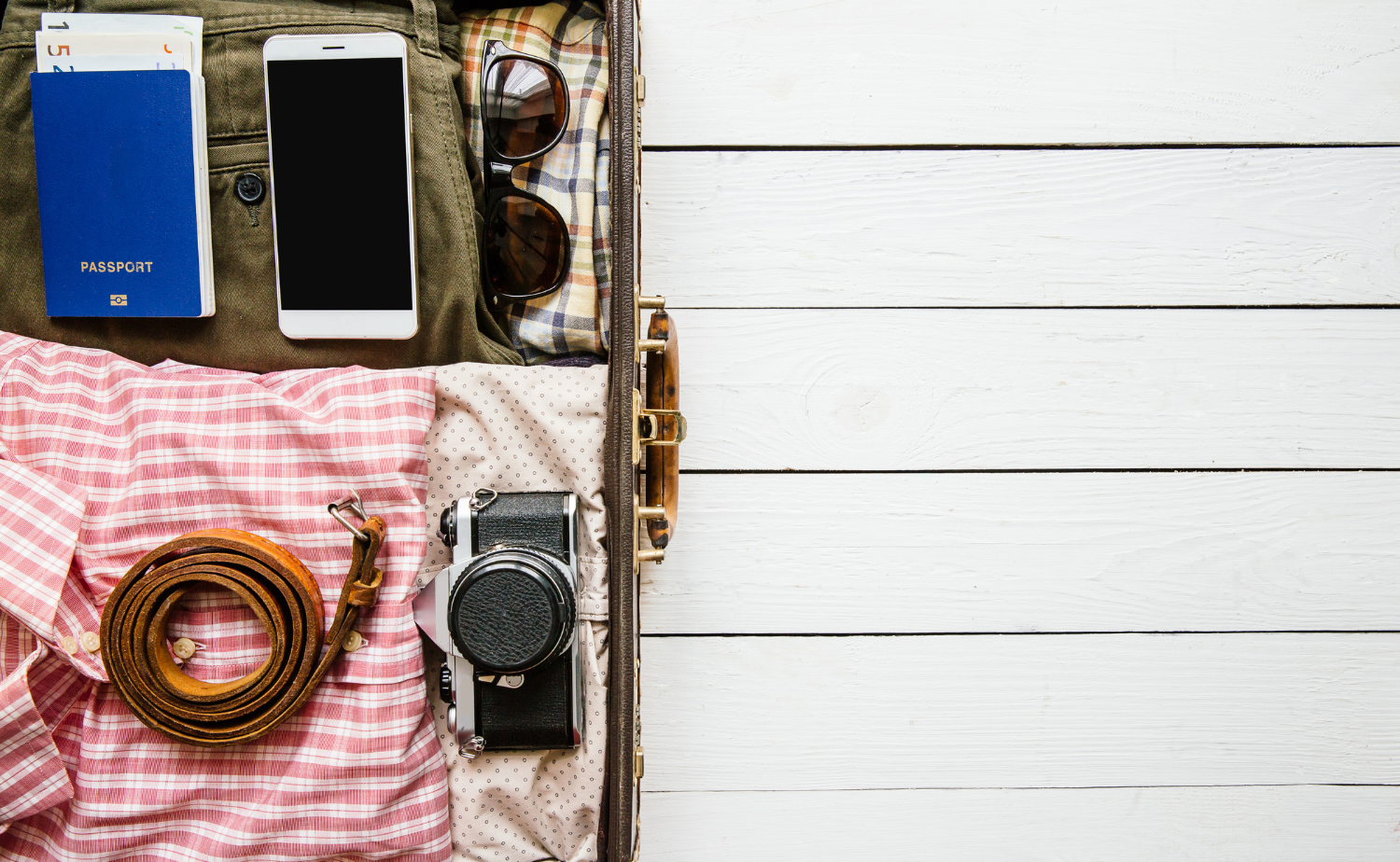 A flatlay of travel essentials including a passport, phone, sunglasses, belt, clothing, and a vintage camera