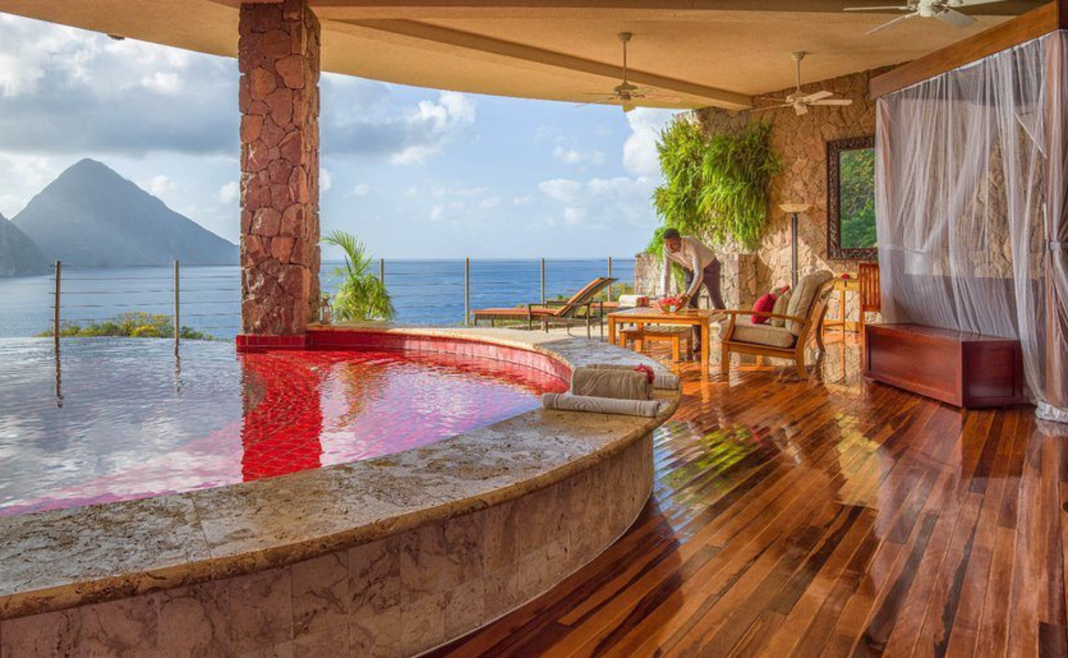 An open-air luxury suite at Jade Mountain overlooks the Caribbean Sea, complete with a red-tiled infinity pool and elegant furnishings framed by stone columns and lush greenery.