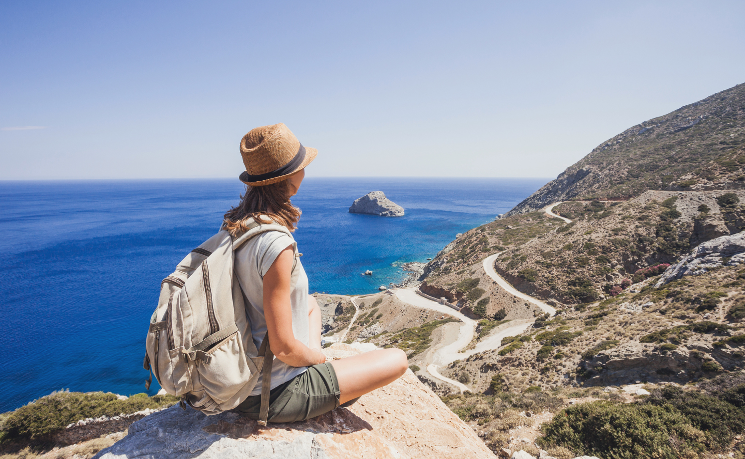 A traveler sitting on a cliff overlooking the Amalfi Coast with winding roads and bright blue Mediterranean water below.