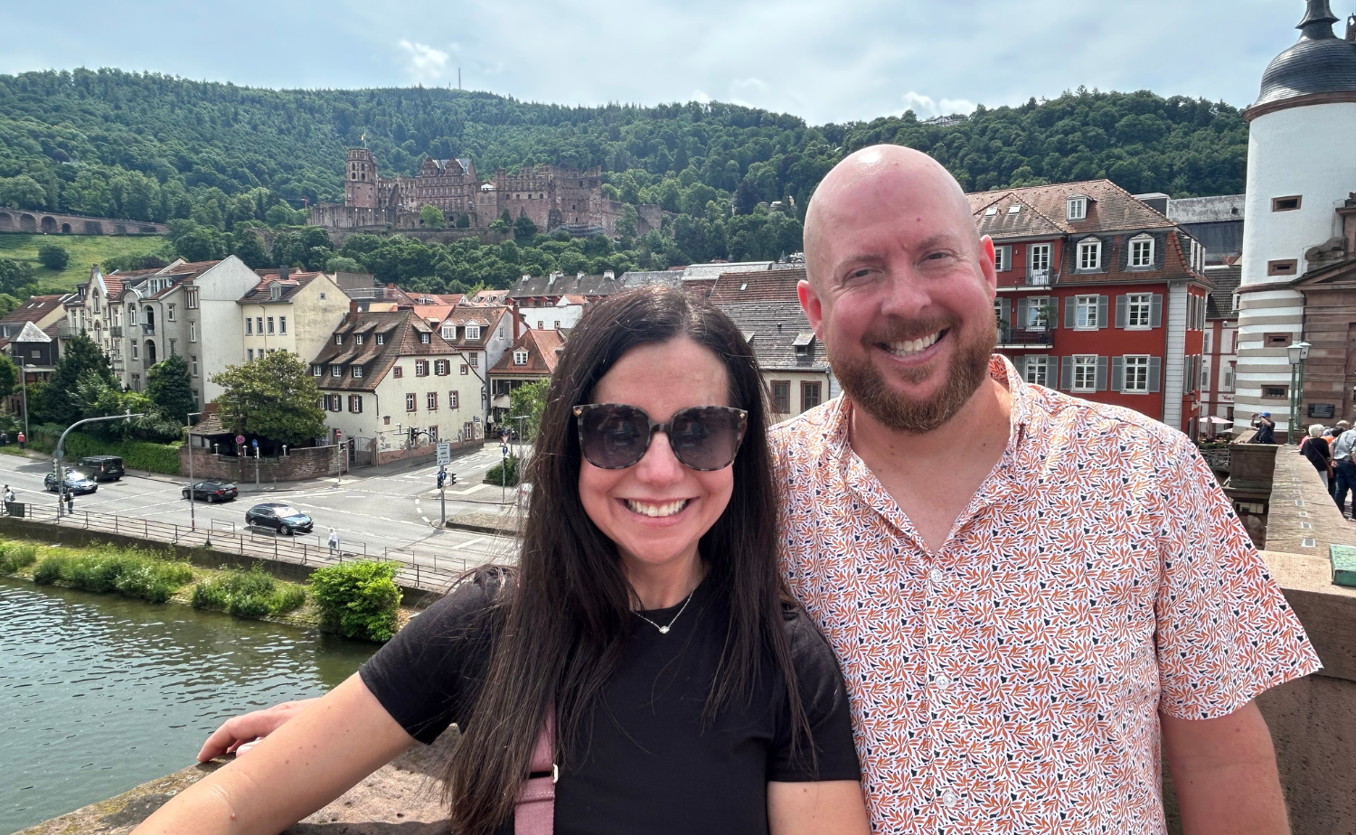 Laura and Kyle smiling together during a Rhine River cruise stop with Heidelberg Castle in the background.
