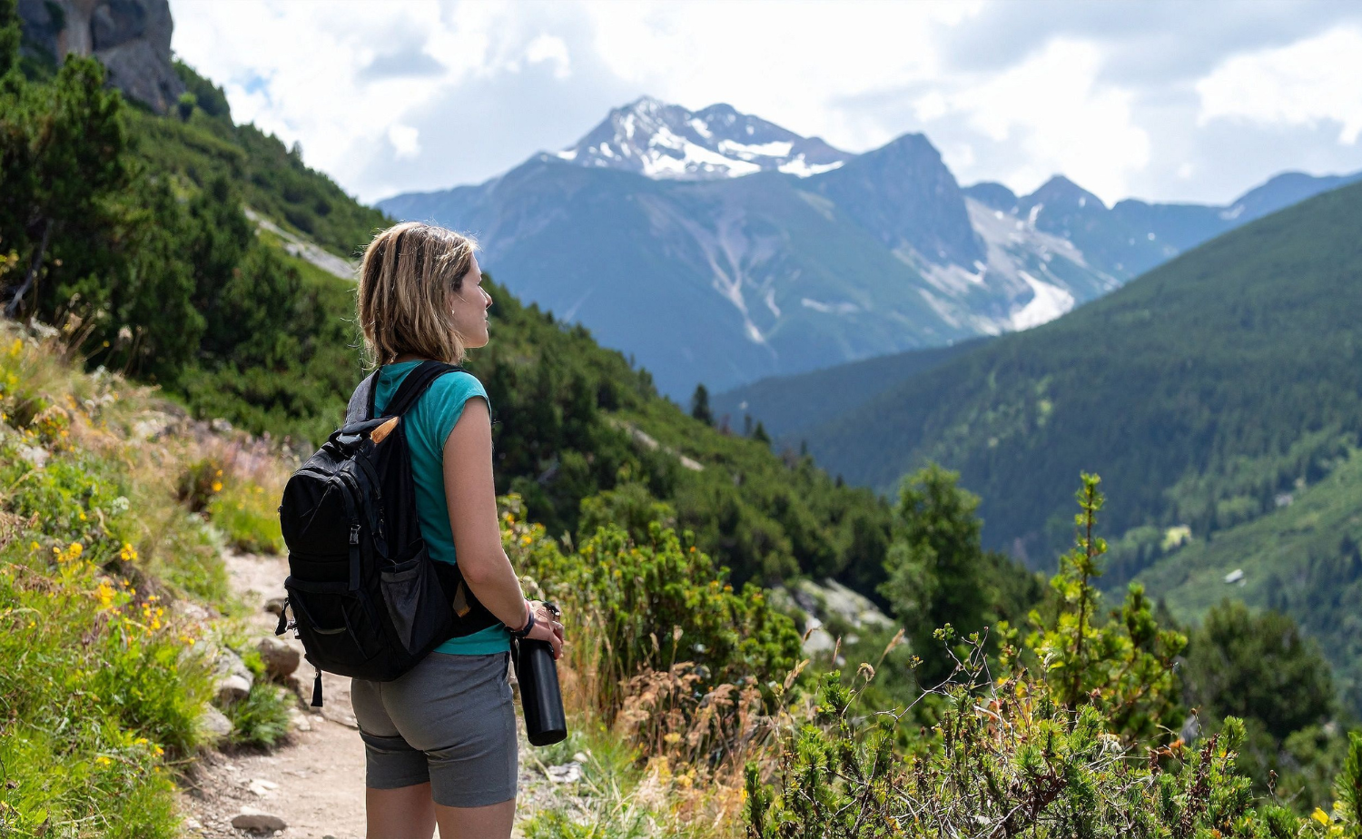 A woman with a backpack pauses on a scenic trail overlooking rugged mountains and forests.