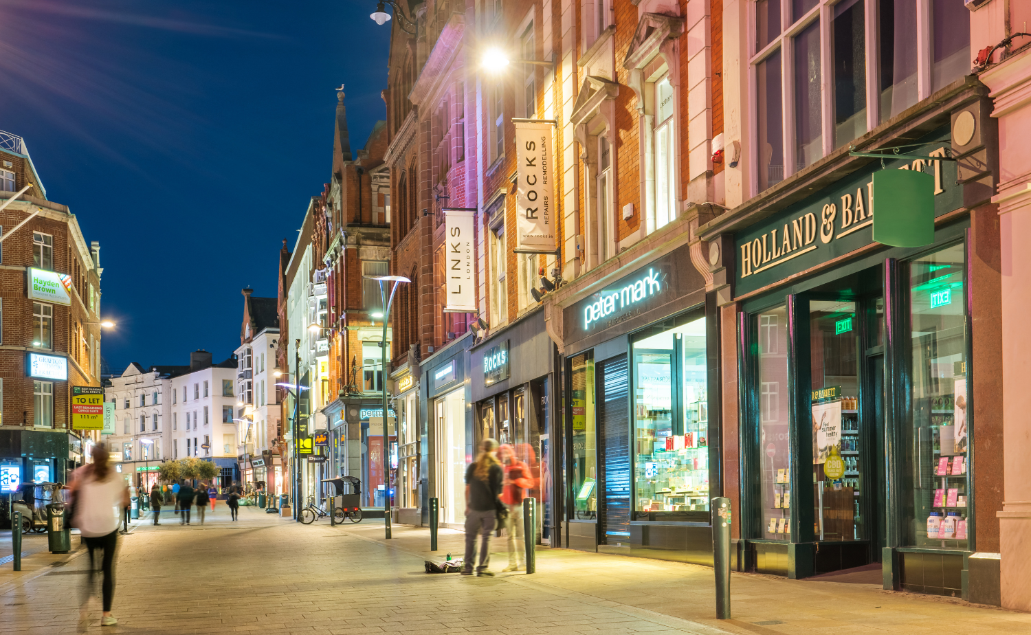 Grafton Street in Dublin at night with illuminated storefronts and people walking along the pedestrian street.