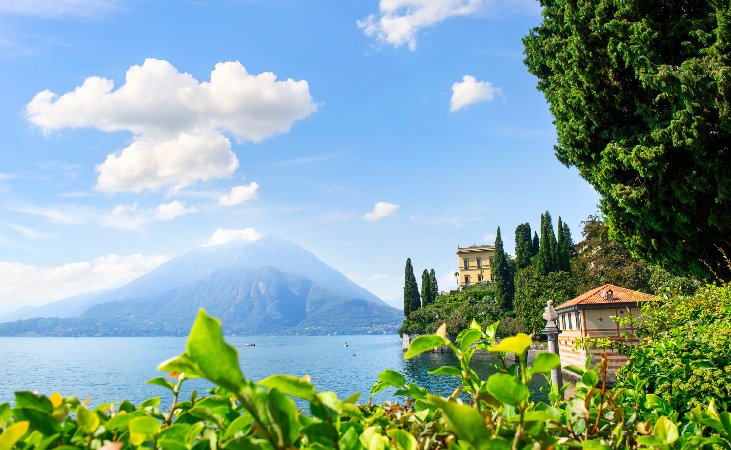 A lakeside villa along Lake Como with mountains rising in the background and lush greenery in the foreground.