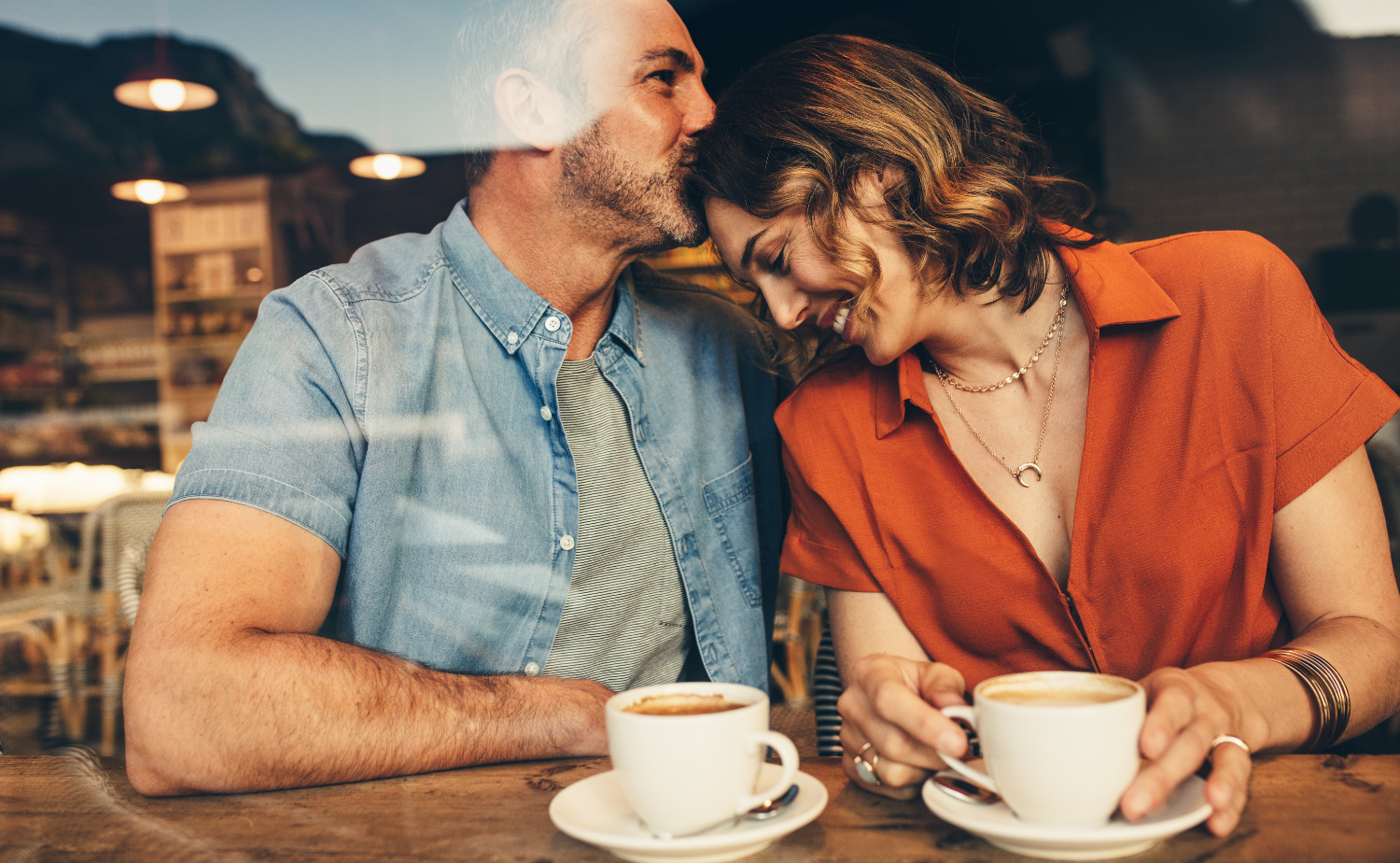 A couple shares a quiet moment over coffee at a cozy café, leaning in close and smiling.