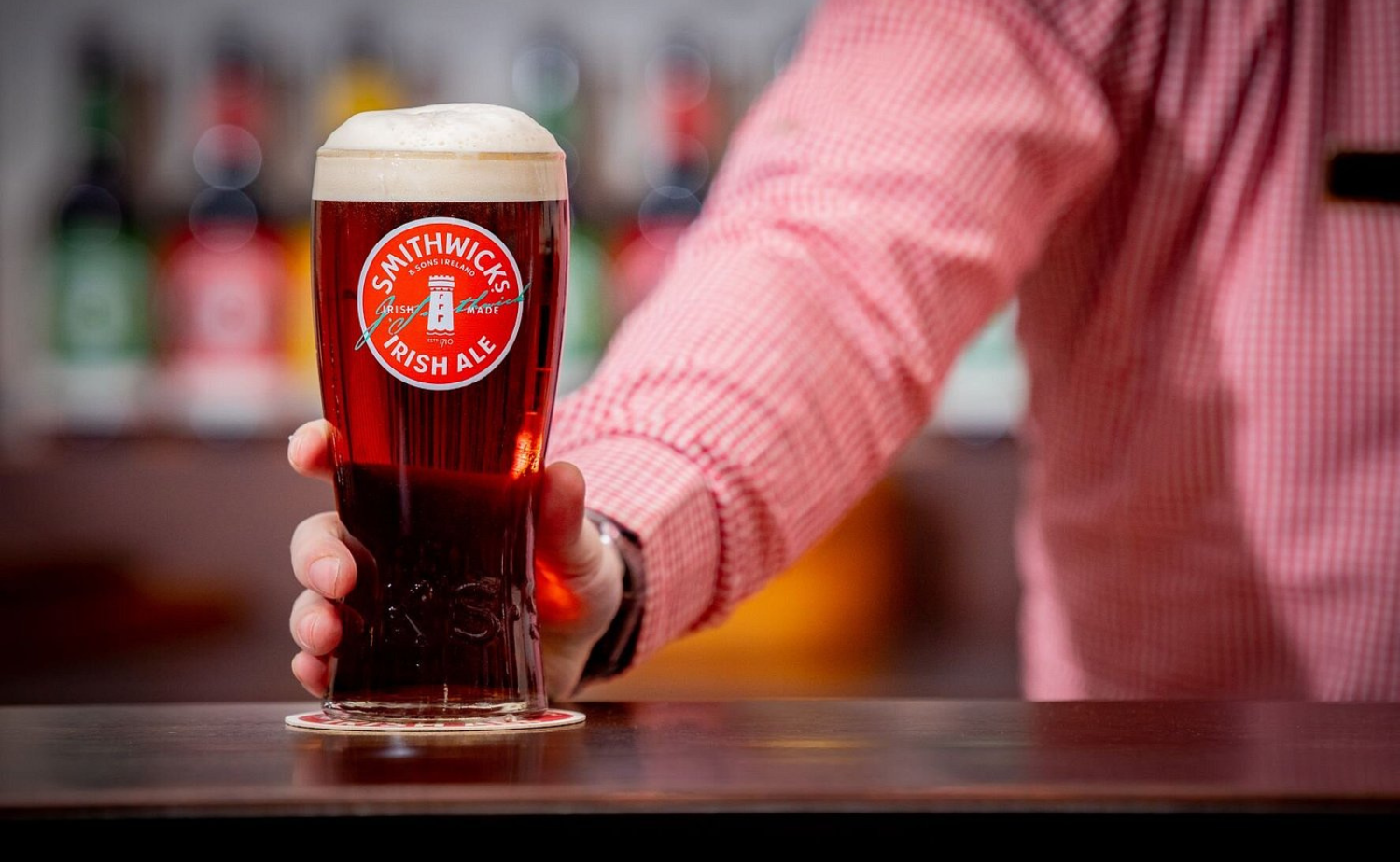 A freshly poured pint of Smithwick’s Irish Ale resting on a bar with bottles blurred in the background.