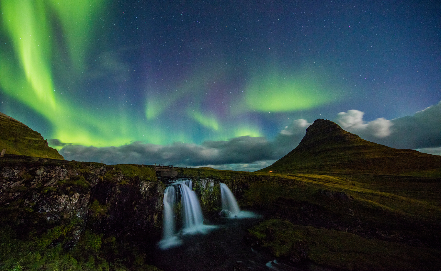 Curtains of green Northern Lights stretch across the sky above Kirkjufell mountain and waterfalls, highlighting Iceland’s iconic natural beauty.
