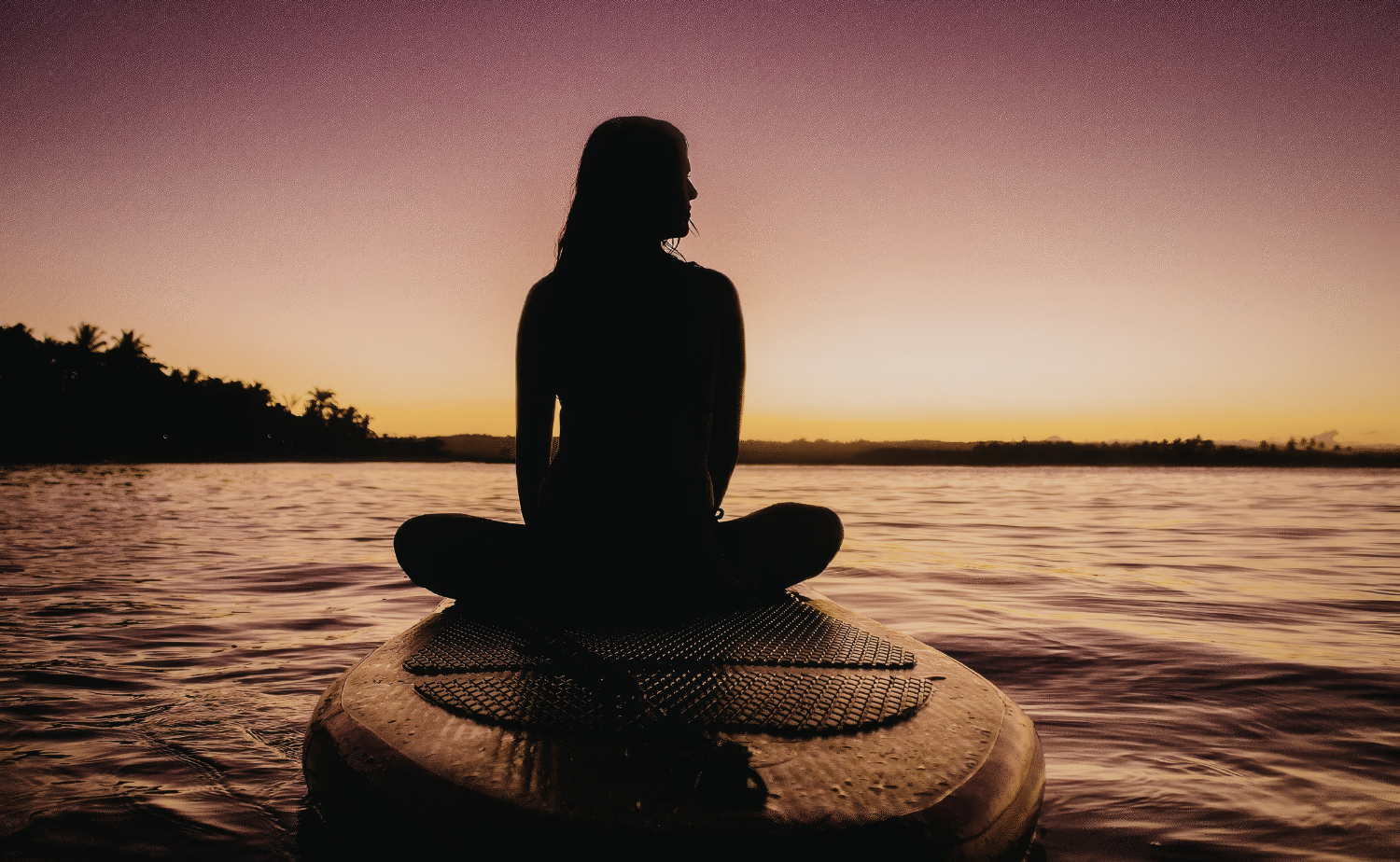 A woman sitting on a paddleboard at sunset, silhouetted against calm island waters.