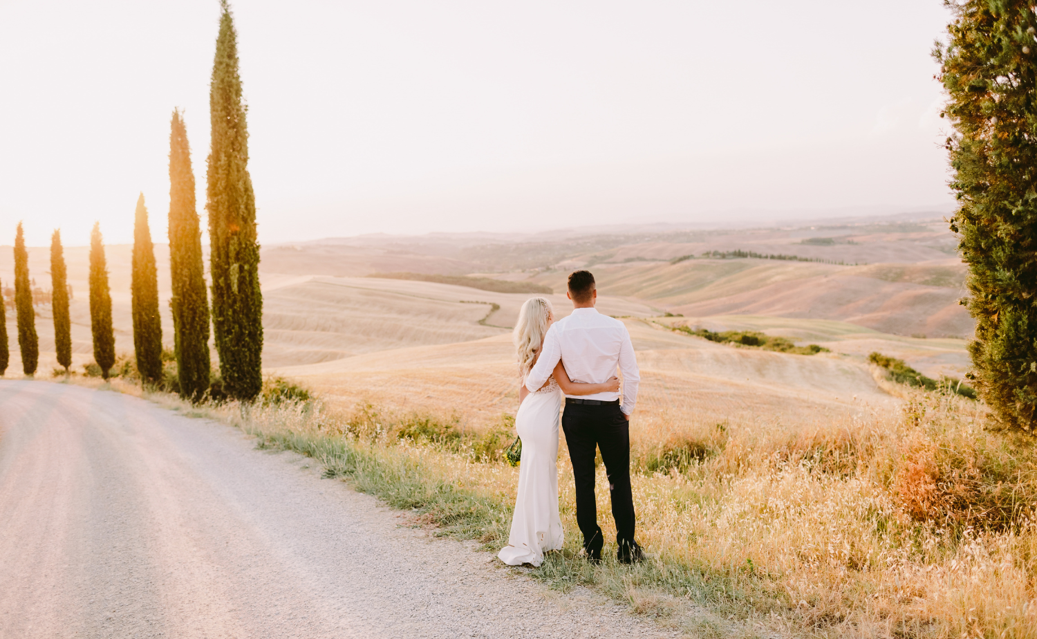A couple stands together along a quiet cypress-lined road overlooking the rolling hills of Tuscany at sunset.