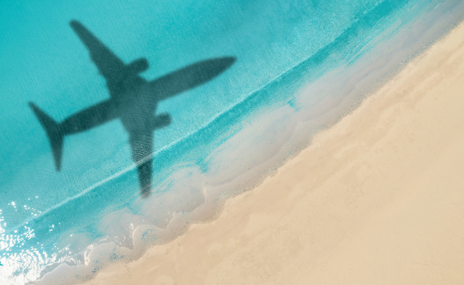 The shadow of an airplane flying over turquoise water and white sand.