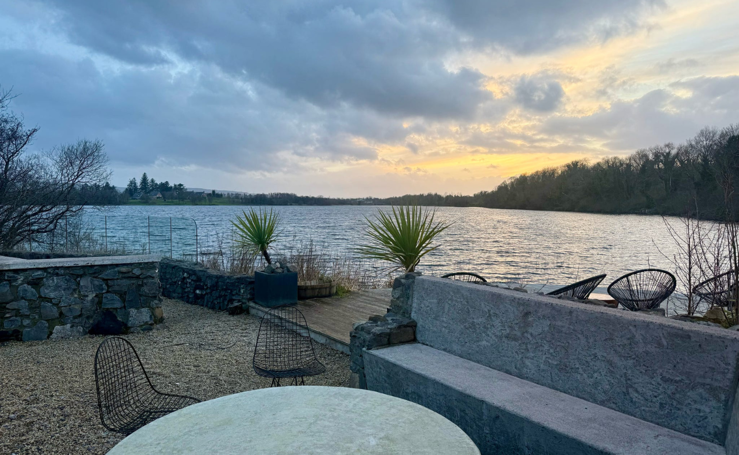 Sunset view over the lake at Finn Lough, with outdoor seating, stone walls, and soft evening light reflecting on the water.