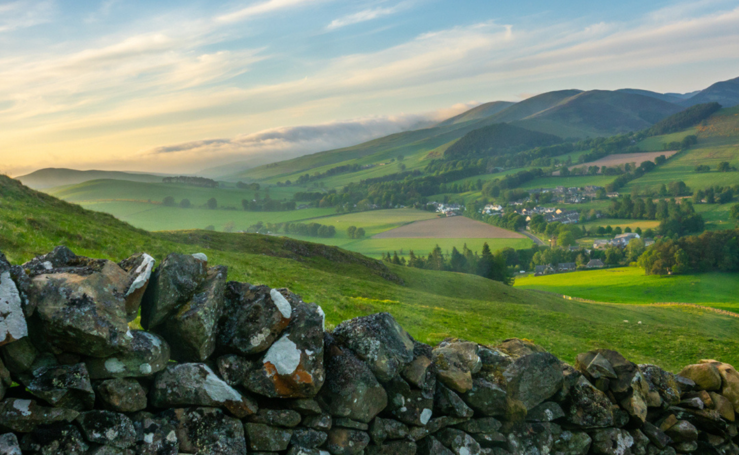 Rolling green hills and patchwork farmland in the Irish countryside under a soft blue sky.
