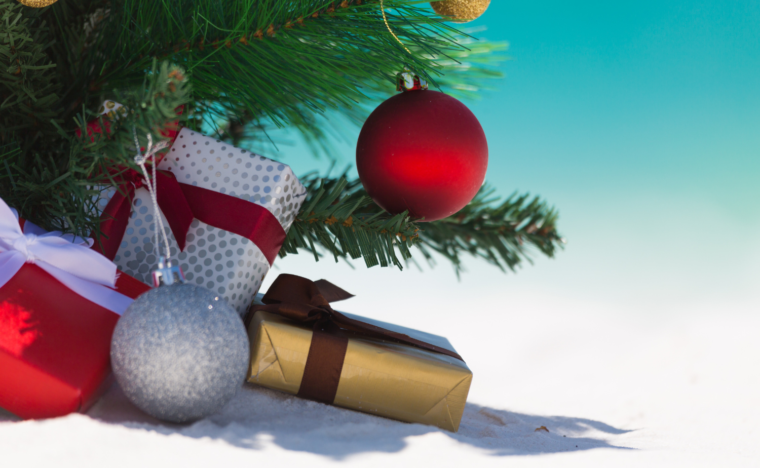 Wrapped Christmas presents and ornaments rest beneath a festive tree on a white sandy beach with turquoise water in the background
