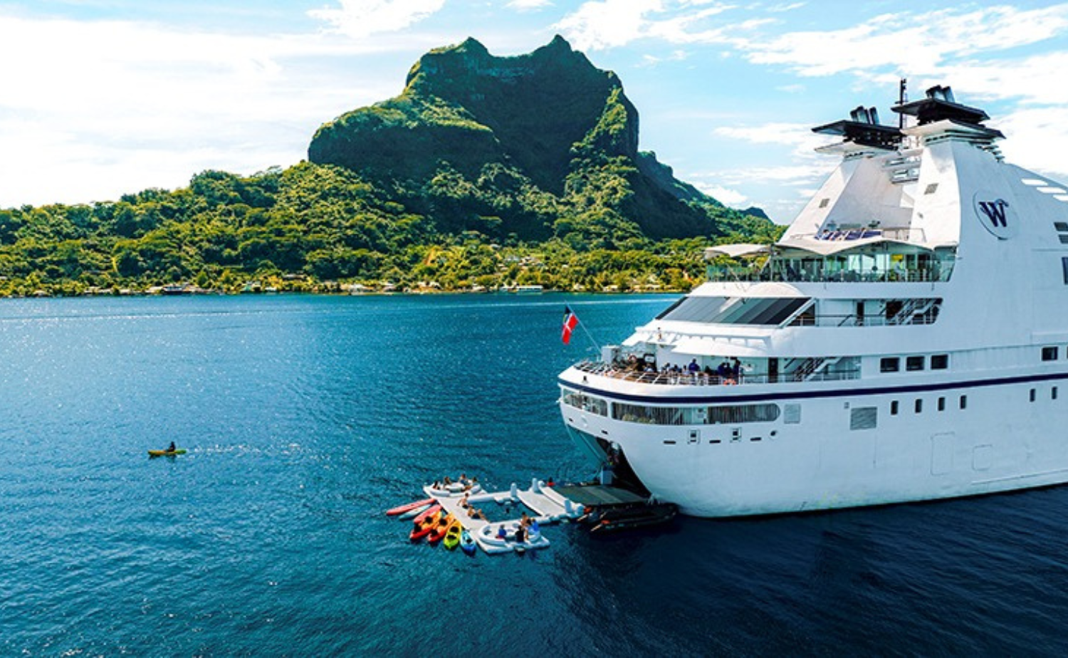 A Windstar yacht anchored in a turquoise bay with lush green mountains rising in the background, featuring kayaks and water sports launched from the ship’s marina—showcasing luxury small-ship cruising in paradise.