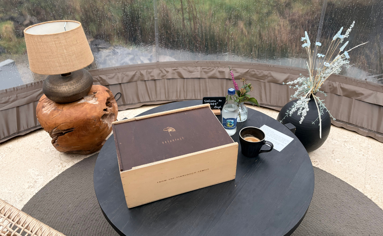 Breakfast delivered inside a Premium River Bubble at Finn Lough, with coffee, water, flowers, and a wooden breakfast box set on a table beside the bubble’s panoramic nature views.