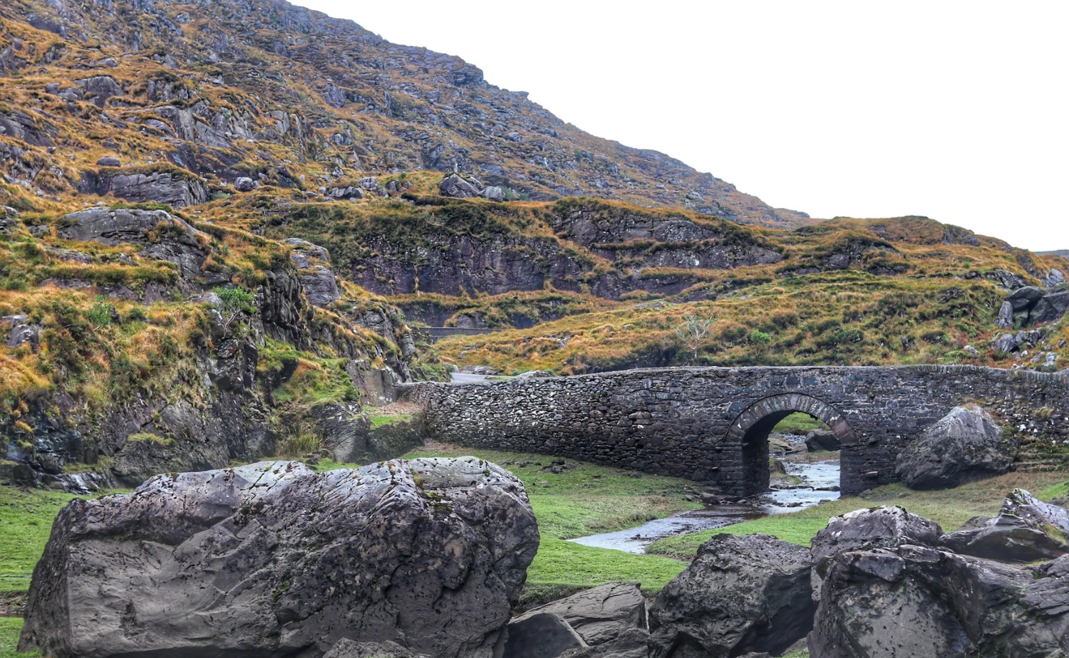 A small stone bridge crossing a stream in the rocky Irish countryside surrounded by hills.