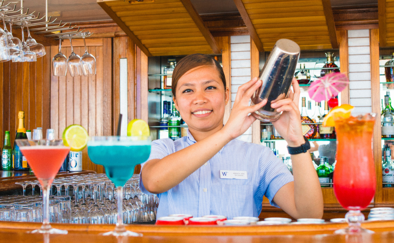 A smiling bartender aboard the Wind Surf cruise ship prepares a cocktail behind a colorful bar.