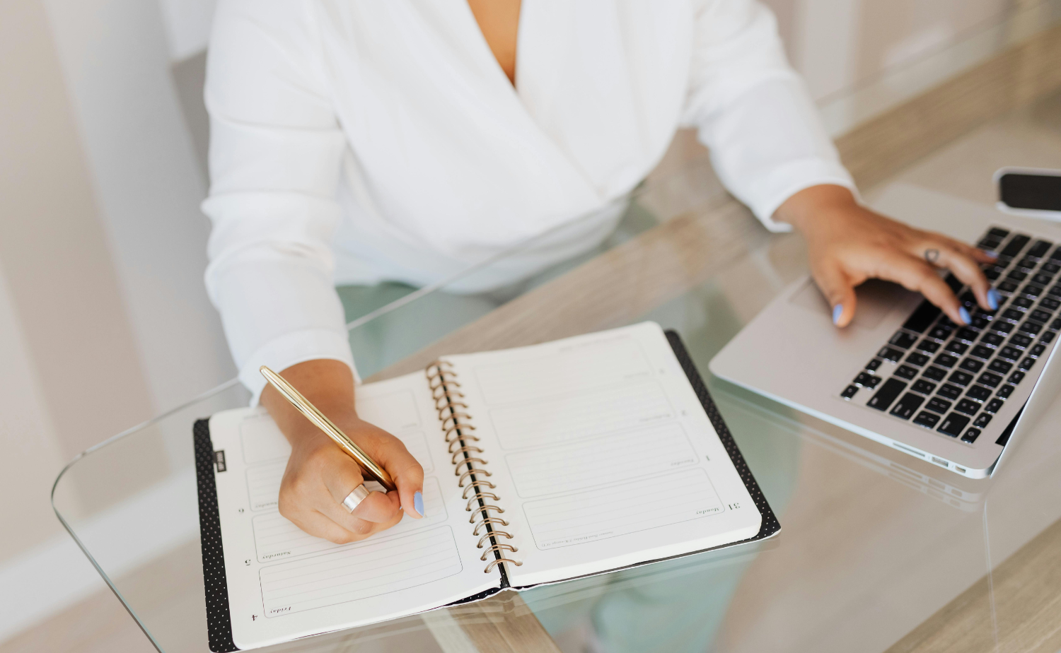 A person writes in an open planner while working on a laptop at a glass desk.