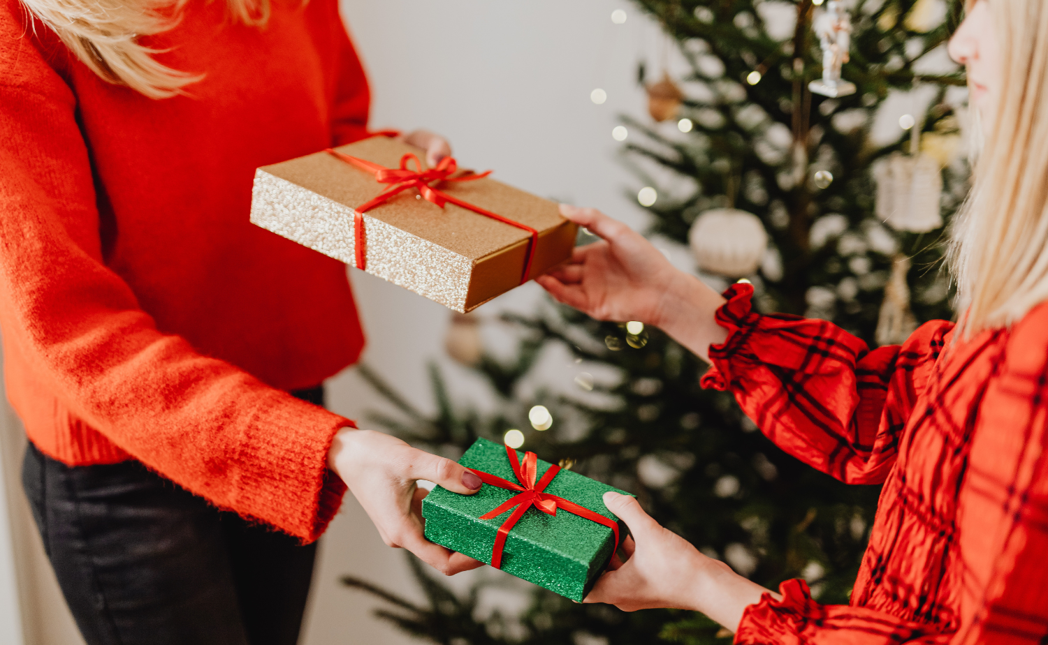 Two people wearing cozy sweaters exchange wrapped gifts in front of a Christmas tree with warm lights in the background