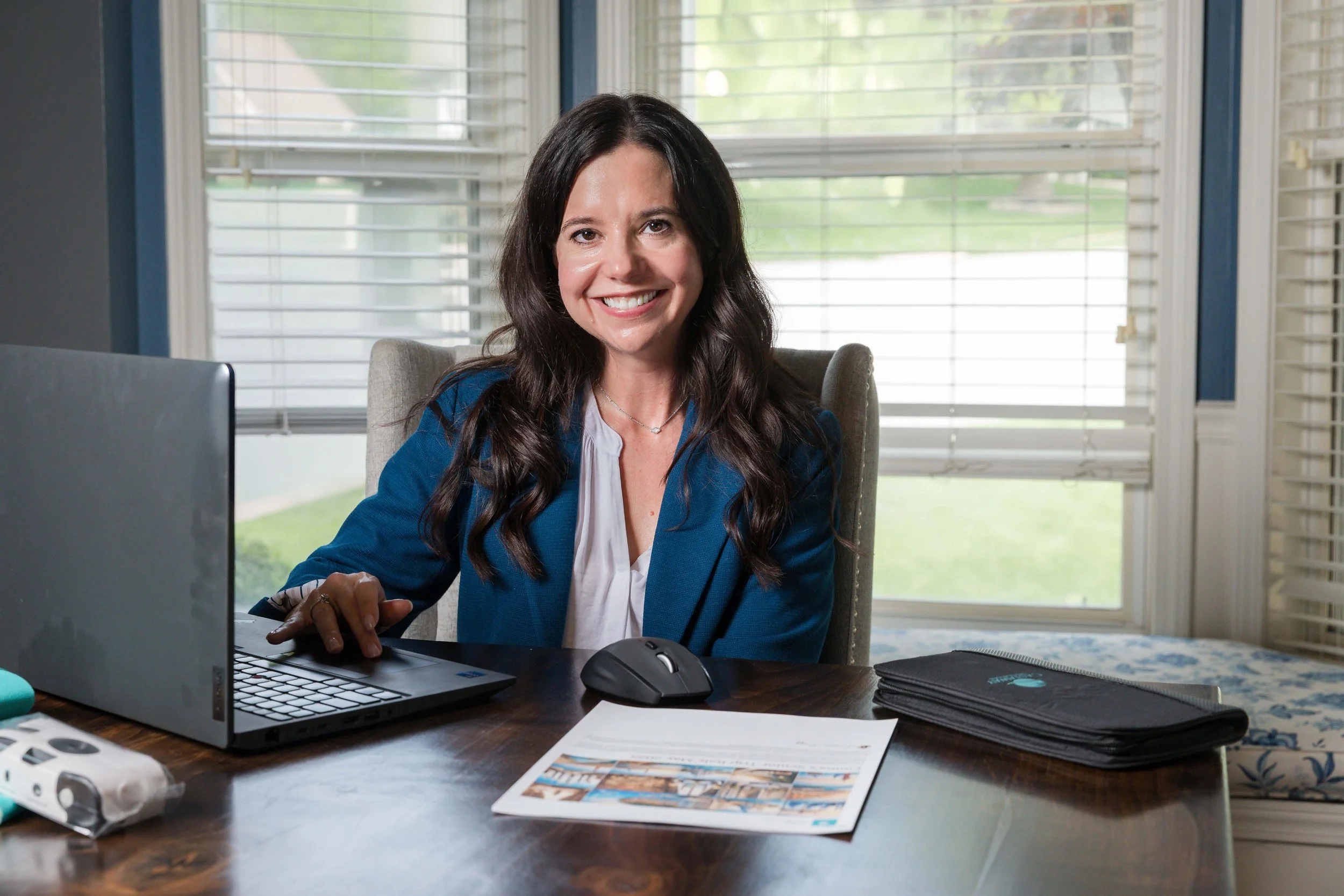 A woman sits at a wooden desk with a laptop and printed itinerary, smiling at the camera.