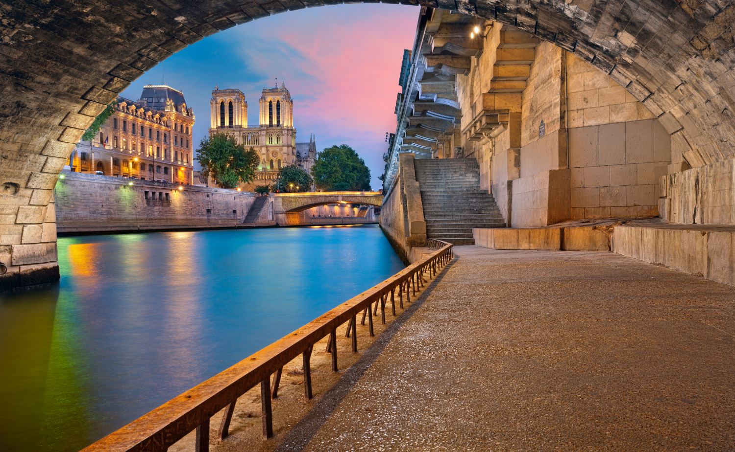 View of the Seine River at dusk with Notre Dame Cathedral framed beneath a stone bridge in Paris.