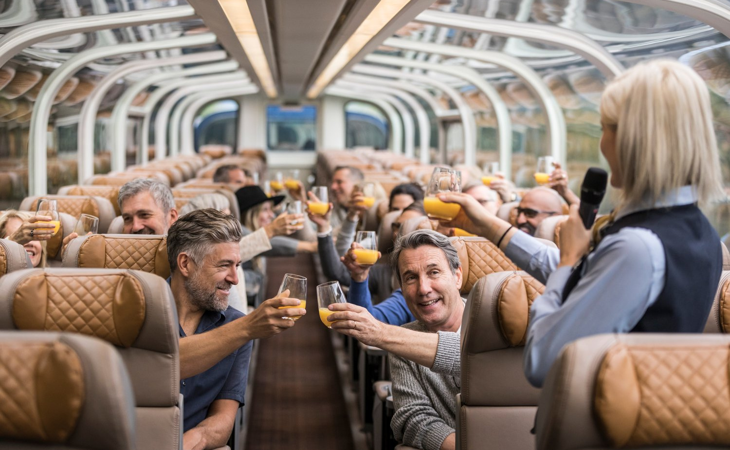 Guests raising glasses for a toast inside the Rocky Mountaineer GoldLeaf dome car with a host speaking at the front.