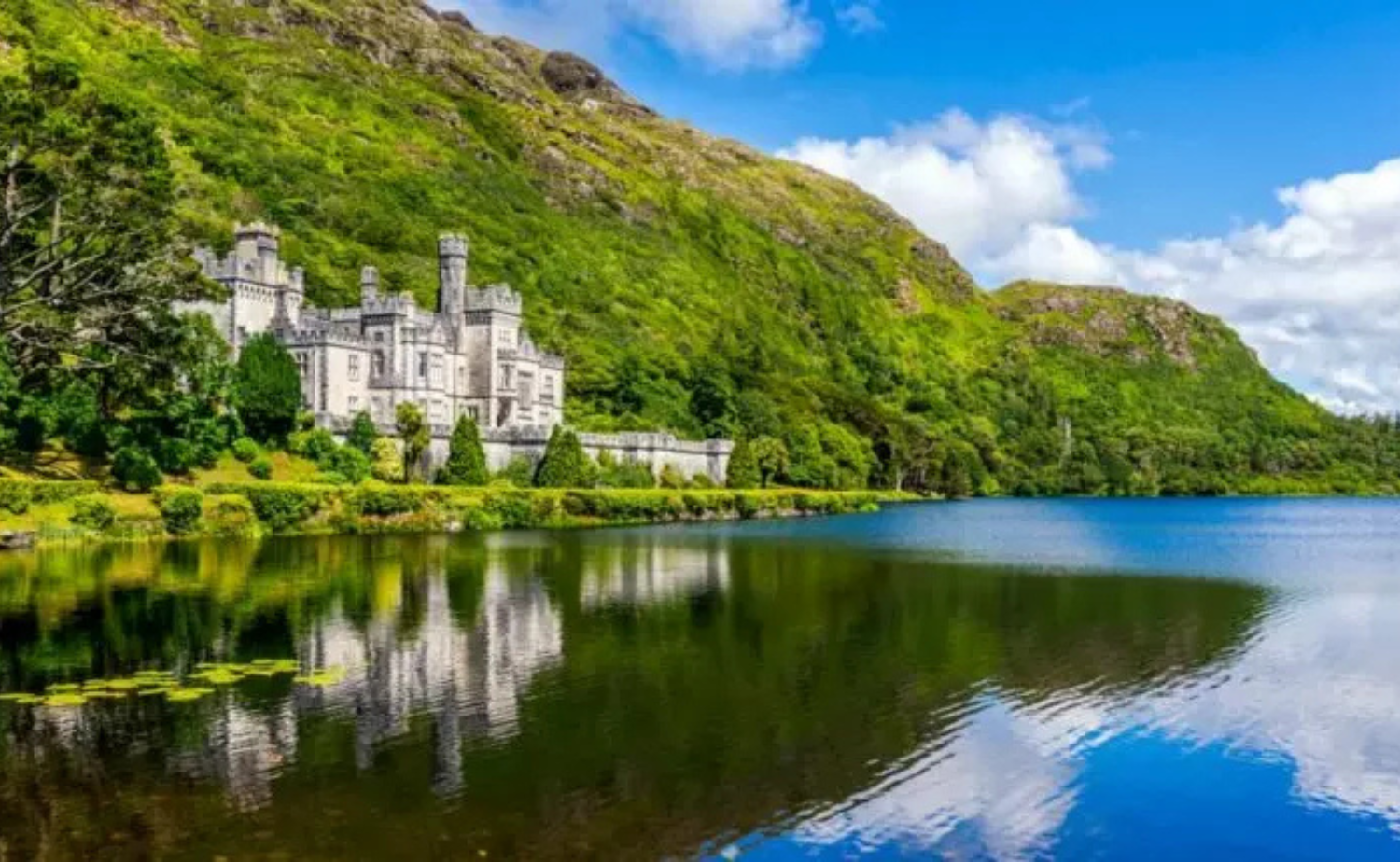 A panoramic view of the Irish countryside with a castle beside a lake and rolling green hills reflected in the water.