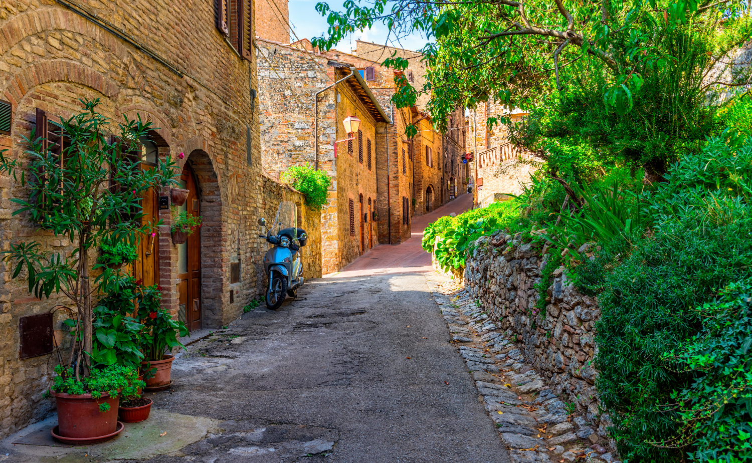 A quiet stone street in a Tuscan village with rustic buildings, greenery, and a parked scooter along the path.