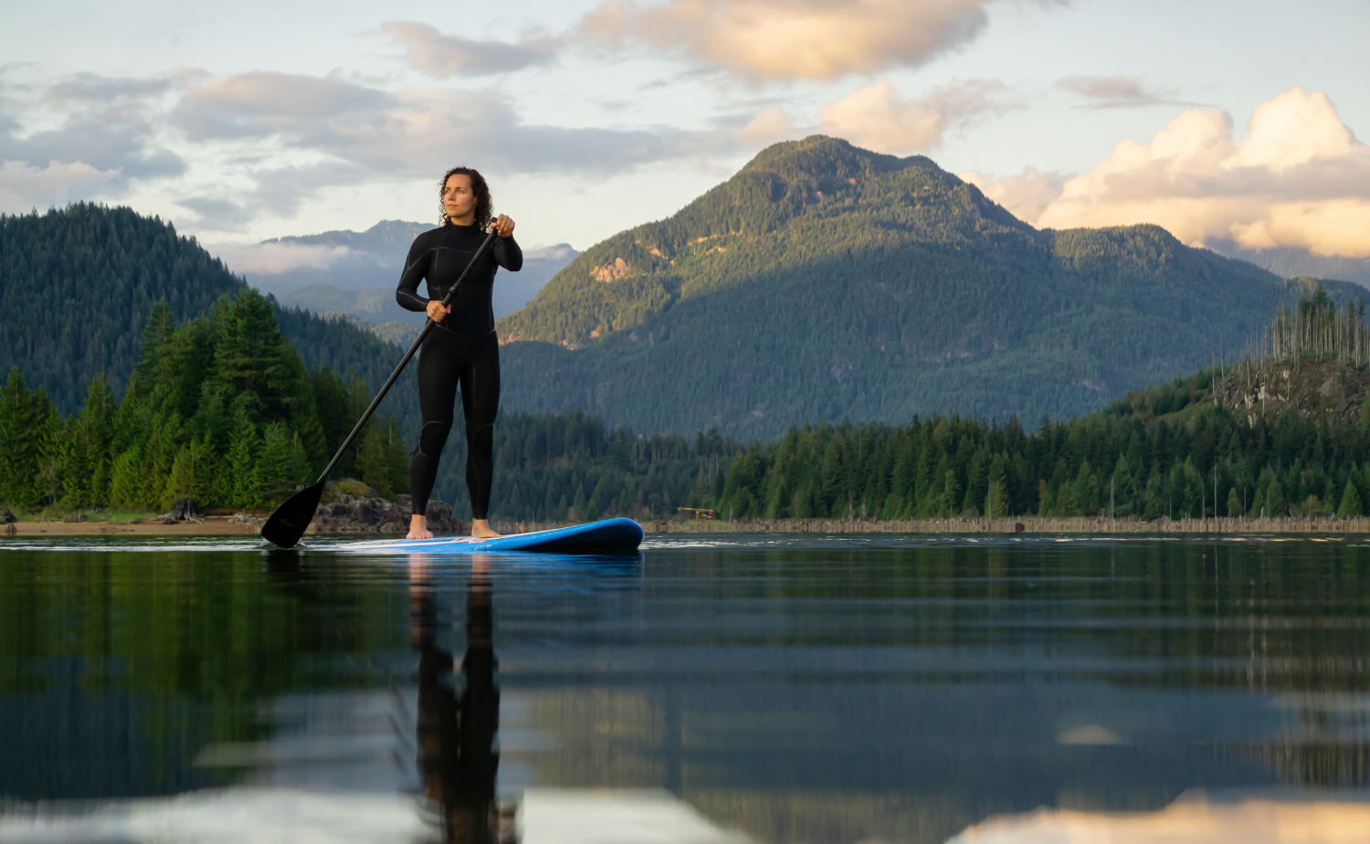 Woman paddleboarding on calm reflective water surrounded by forested mountains, representing the relaxed and unhurried rhythm of island travel.