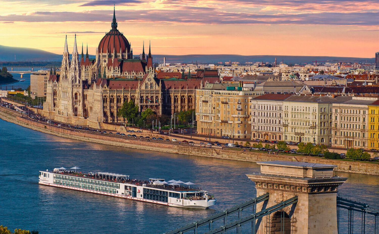 River cruise ship sailing along the Danube River in Budapest with the Hungarian Parliament Building at sunset.