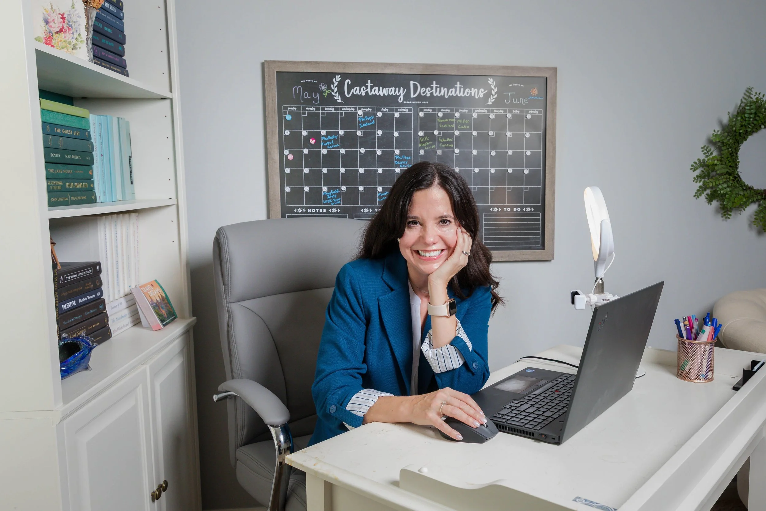 A woman seated at her desk smiling while working on a laptop with a travel planning calendar behind her.