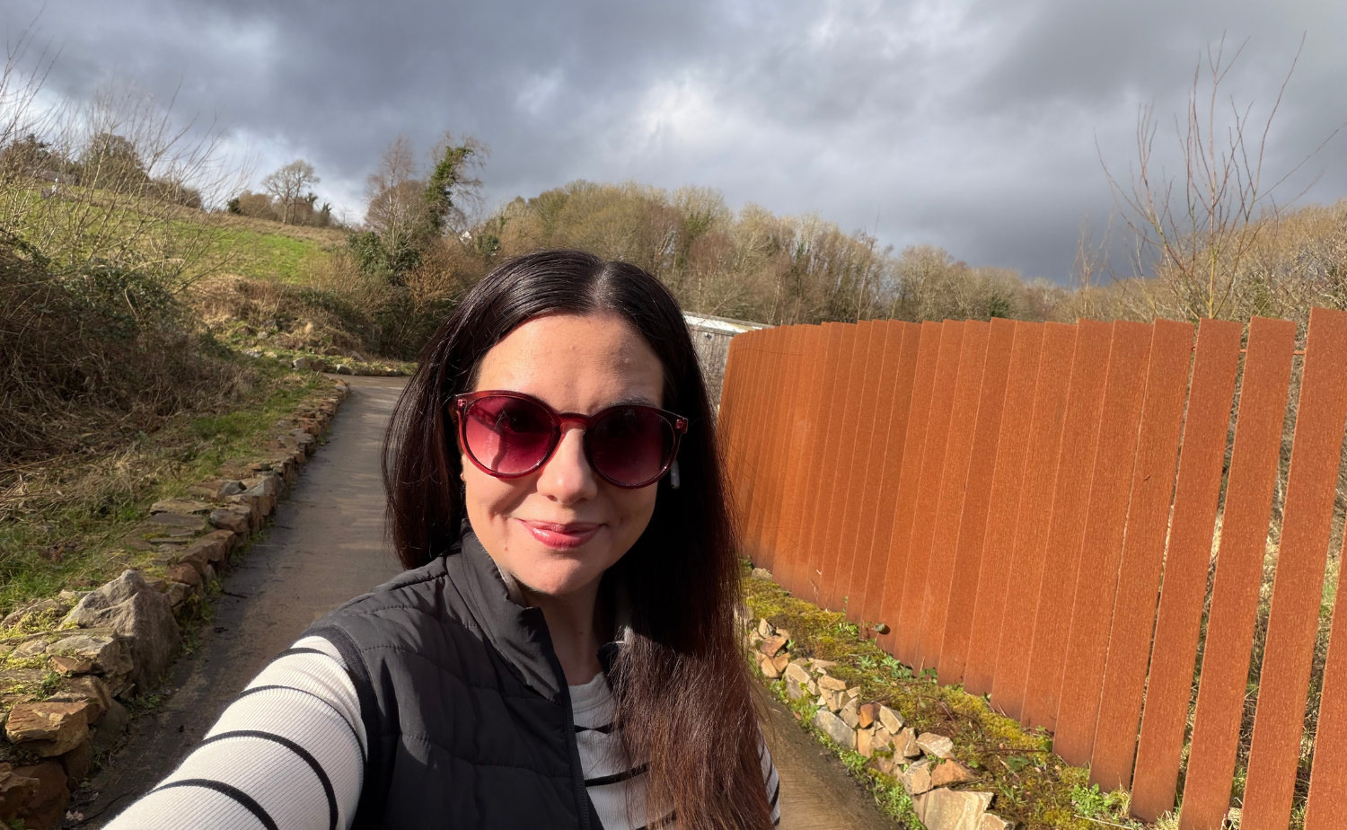 Laura smiling on a walking path at Finn Lough in Northern Ireland, with wooded countryside and modern rust-colored fencing in the background.