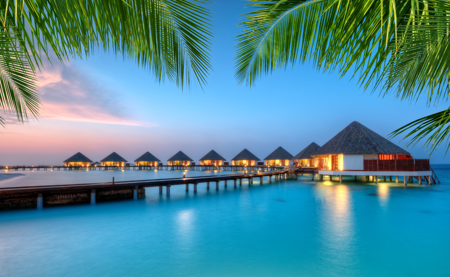 Sunset overwater villas surrounded by turquoise water and palm trees at a luxury resort in the Maldives.