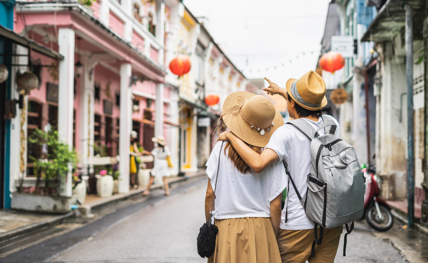 A couple wearing sun hats walks arm in arm down a colorful historic street, pointing out sights together.