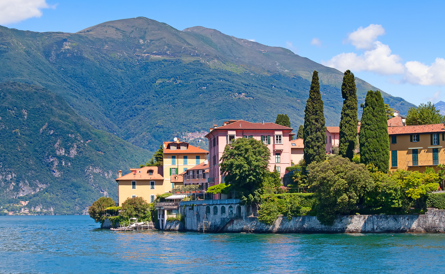 A scenic view of Lake Como with colorful villas, cypress trees, and mountains rising in the background.