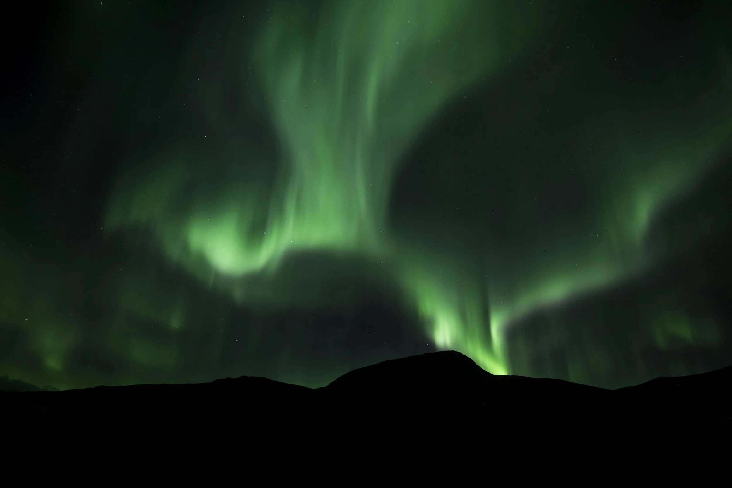 Northern Lights in green hues swirling in the night sky over dark silhouette of mountains.