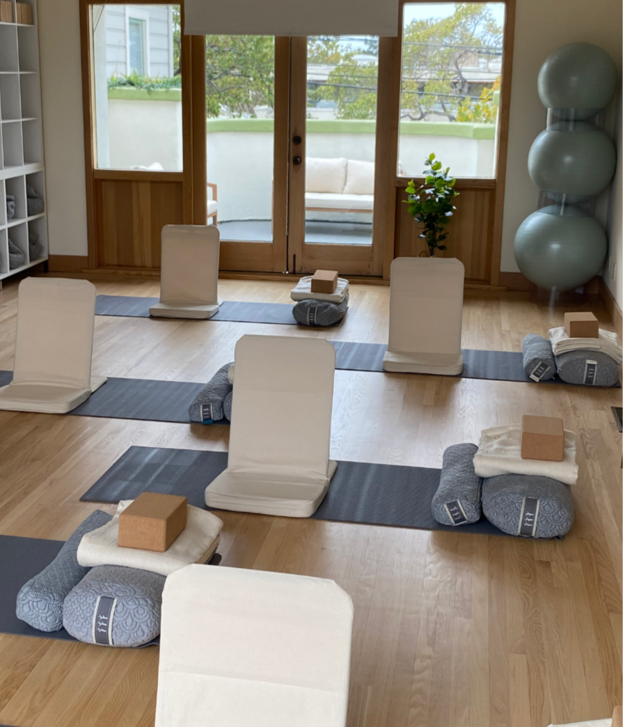 The Brilliant Births studio is set up as a yoga studio with mats, bolsters, blocks, and white floor chairs. In the background, large glass doors lead to a small outdoor balcony with seating.