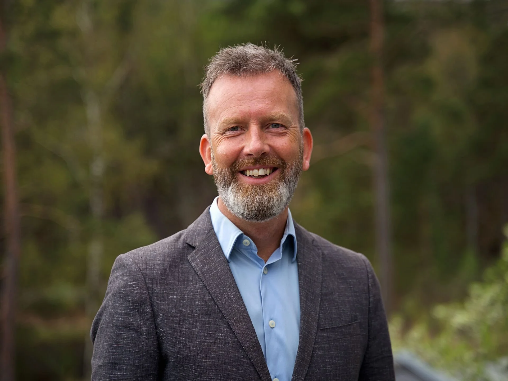 A smiling man with a beard and short hair, wearing a gray blazer and a light blue dress shirt, standing outdoors in front of a blurred forest background.