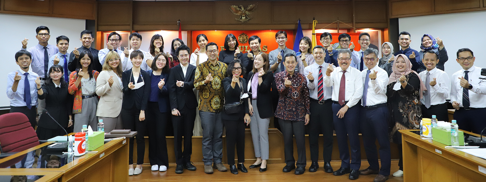 Group of approximately 30 men and women gathered in a conference room, smiling, making a gesture with their hands, with flags and symbols on the wall behind them.