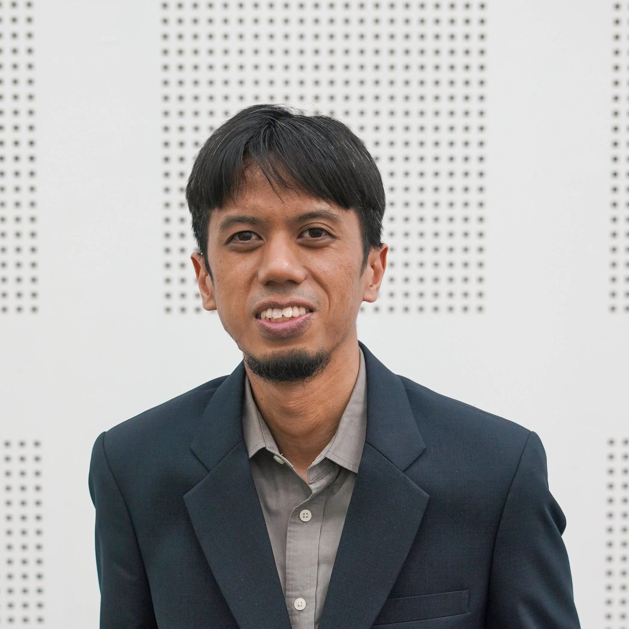 A man with short black hair, a goatee, and wearing a dark suit jacket over a gray shirt, posing in front of a white wall with black perforated patterns.