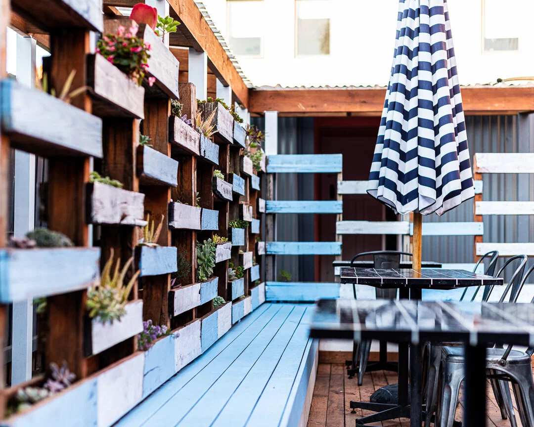 Outdoor patio with wooden and painted pallet wall adorned with small potted plants, striped blue and white umbrella over black metal tables and chairs.